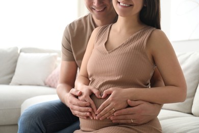 Photo of Pregnant young woman making heart with hands on belly and husband near her at home, closeup