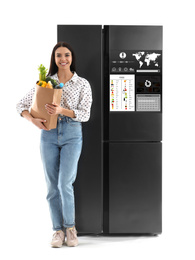 Young woman with bag of groceries near smart refrigerator on white background