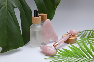 Photo of Gua sha stone, face roller, cosmetic products and green leaves on light background