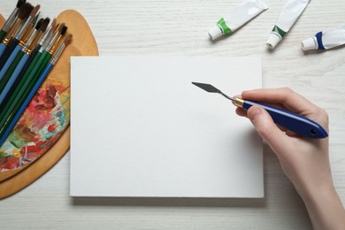 Man with spatula and blank canvas at white wooden table, top view