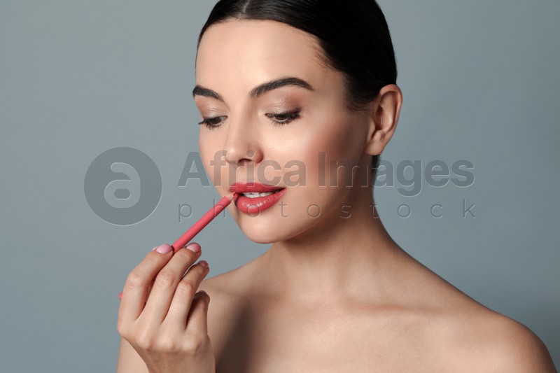 Pretty young woman applying beautiful pink lip pencil on grey background