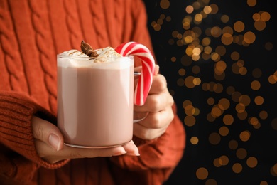 Woman holding glass cup of tasty cocoa with marshmallows and Christmas candy cane against blurred festive lights, closeup. Space for text