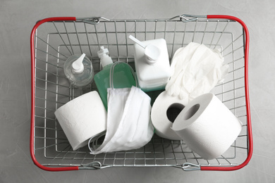 Shopping basket with antiseptics, toilet paper, gloves and masks on light grey stone table, top view. Panic caused by virus