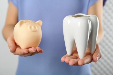 Woman holding ceramic model of tooth and piggy bank on light background, closeup. Expensive treatment