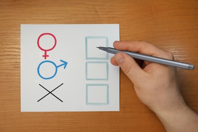 Photo of Gender equality. Man holding pen over sheet of paper with cross mark, male and female symbols at wooden table, top view