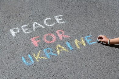 Photo of Girl writing Peace For Ukraine with colorful chalks on asphalt outdoors, closeup