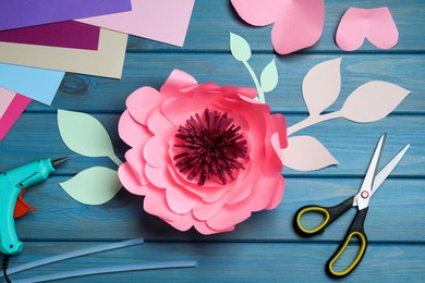Photo of Beautiful paper flower, scissors and hot glue gun on blue wooden background, flat lay. Master class in handmade craft