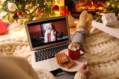 Photo of MYKOLAIV, UKRAINE - DECEMBER 23, 2020: Woman with cocoa and wafers watching The Christmas Chronicles movie on laptop near fireplace at home, closeup. Cozy winter holidays atmosphere