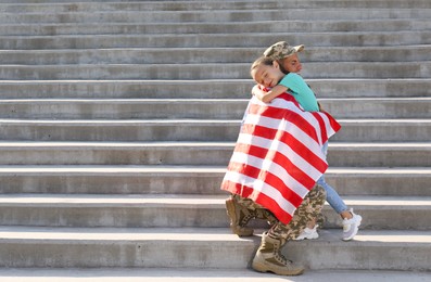 Soldier with flag of USA and his little daughter hugging outdoors, space for text
