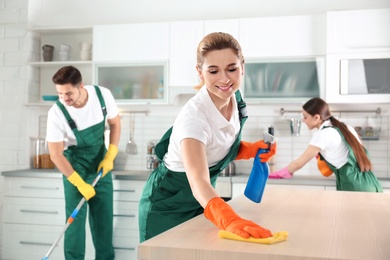 Woman using rag and sprayer for cleaning table with colleagues in kitchen