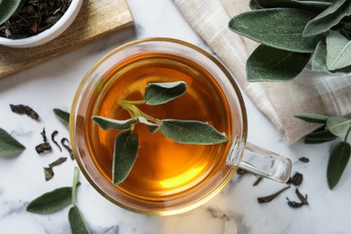 Cup of aromatic sage tea with fresh leaves on white marble table, flat lay