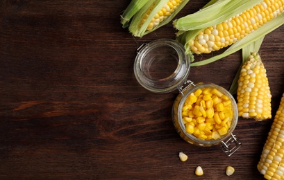 Tasty sweet corn kernels in jar and fresh cobs on wooden table, flat lay. Space for text