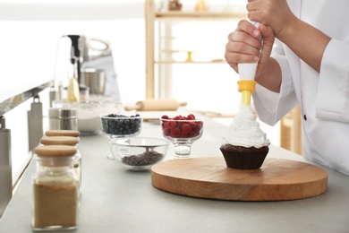 Young female pastry chef decorating cupcake with cream at table in kitchen, closeup