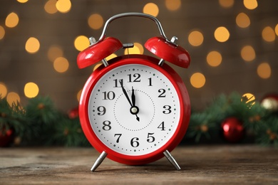 Alarm clock with decor on wooden table against blurred Christmas lights, closeup. New Year countdown