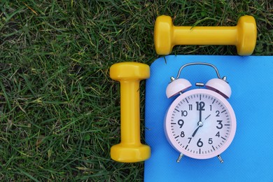 Photo of Alarm clock, dumbbells and fitness mat on green grass, flat lay with space for text. Morning exercise