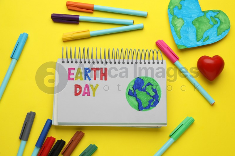 Notebook with phrase Earth Day, heart shaped model of planet and felt pens on yellow background, flat lay