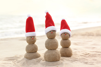 Snowmen made of sand with Santa hats on beach near sea. Christmas vacation