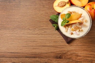 Tasty peach yogurt with granola, mint and pieces of fruit in dessert bowl on wooden table, flat lay. Space for text