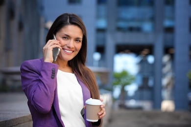 Beautiful businesswoman with cup of coffee talking on phone outdoors. Space for text