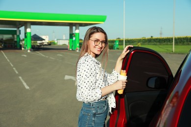 Beautiful young woman with hot dog opening car door at gas station. Space for text