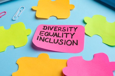 Sticky note with words Diversity, Equality, Inclusion and paper puzzle pieces on light blue background, closeup