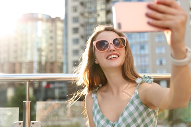 Photo of Beautiful young woman with sunglasses taking selfie outdoors, space for text