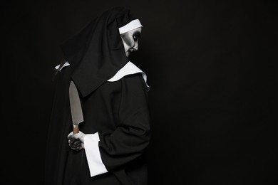 Photo of Scary devilish nun with knife behind back on black background, space for text. Halloween party look