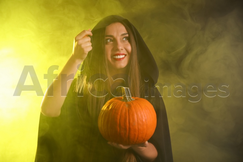 Young woman wearing witch costume with pumpkin in smoke cloud on dark background. Halloween party
