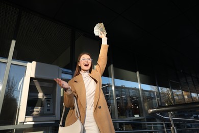 Excited young woman with money near cash machine outdoors
