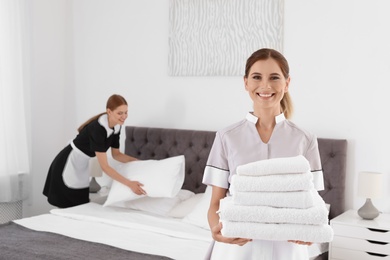 Professional chambermaid holding pile of clean towels in bedroom