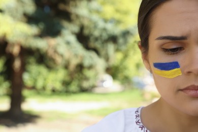 Photo of Sad young woman with drawing of Ukrainian flag on face outdoors, closeup. Space for text