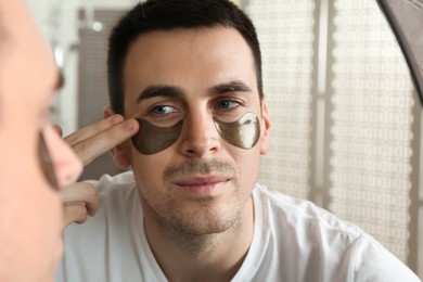 Young man applying under eye patches near mirror at home, closeup