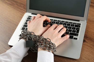 Woman with chained hands typing on laptop at wooden table, closeup. Internet addiction