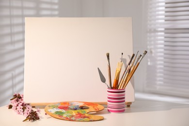 Easel with blank canvas, fresh flowers and art supplies on white table