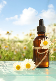 Bottle of chamomile essential oil on light blue wooden table in field