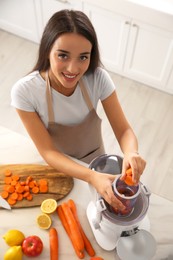 Young woman putting fresh slices of carrot into juicer at table in kitchen, above view
