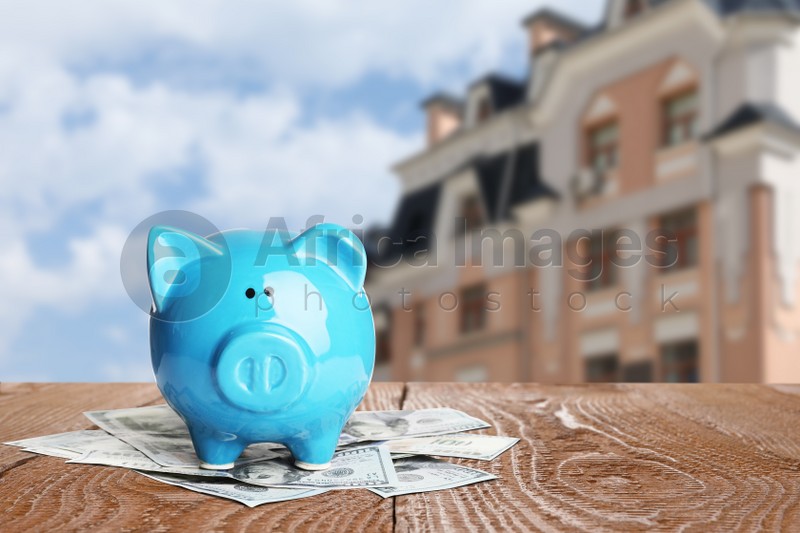 Piggy bank and money on wooden surface and blurred view of beautiful house, space for text. Mortgage concept