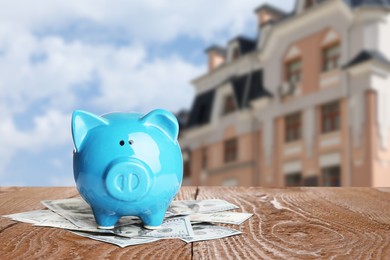 Piggy bank and money on wooden surface and blurred view of beautiful house, space for text. Mortgage concept