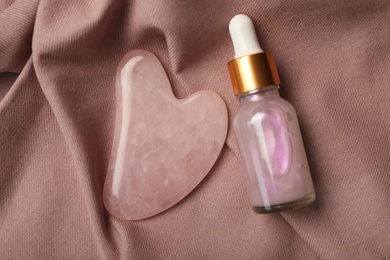 Photo of Rose quartz gua sha tool and skin care product on brown fabric, flat lay