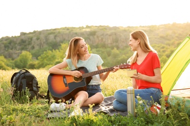 Young women resting with hot drink and guitar near camping tent in wilderness