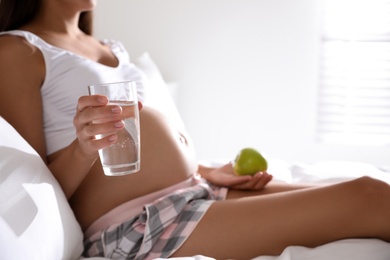 Young pregnant woman with glass of water and apple in bedroom, closeup. Taking care of baby health
