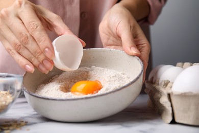Photo of Woman adding egg to bowl with flour at white marble table in kitchen, closeup. Cooking oatmeal dough