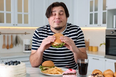 Happy overweight man with tasty burger at table in kitchen