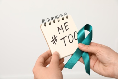 Woman holding notebook with hashtag MeToo and teal awareness ribbon against light background, closeup. Stop sexual assault