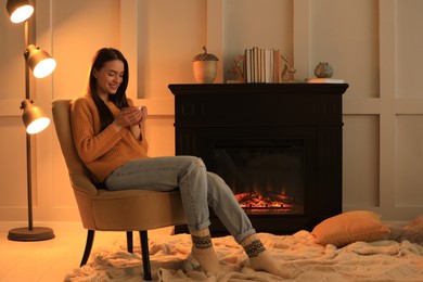 Photo of Young woman with cup of hot drink near fireplace at home. Cozy atmosphere