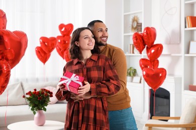 Photo of Happy couple celebrating Valentine's day. Beloved woman with gift box in room decorated with heart shaped air balloons