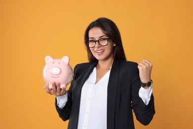 Photo of Happy young woman in eyeglasses with piggy bank on orange background