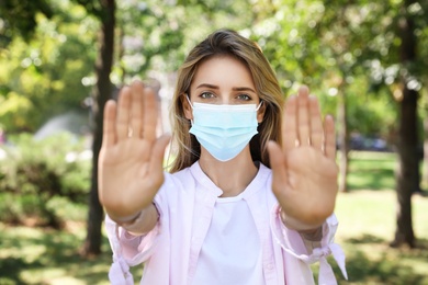 Woman in protective face mask showing stop gesture in park. Prevent spreading of coronavirus