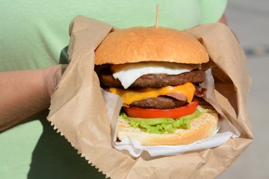 Photo of Woman holding delicious burger in paper wrap on blurred background, closeup