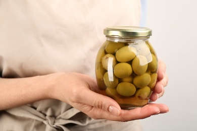 Woman holding glass jar of pickled olives on light background, closeup